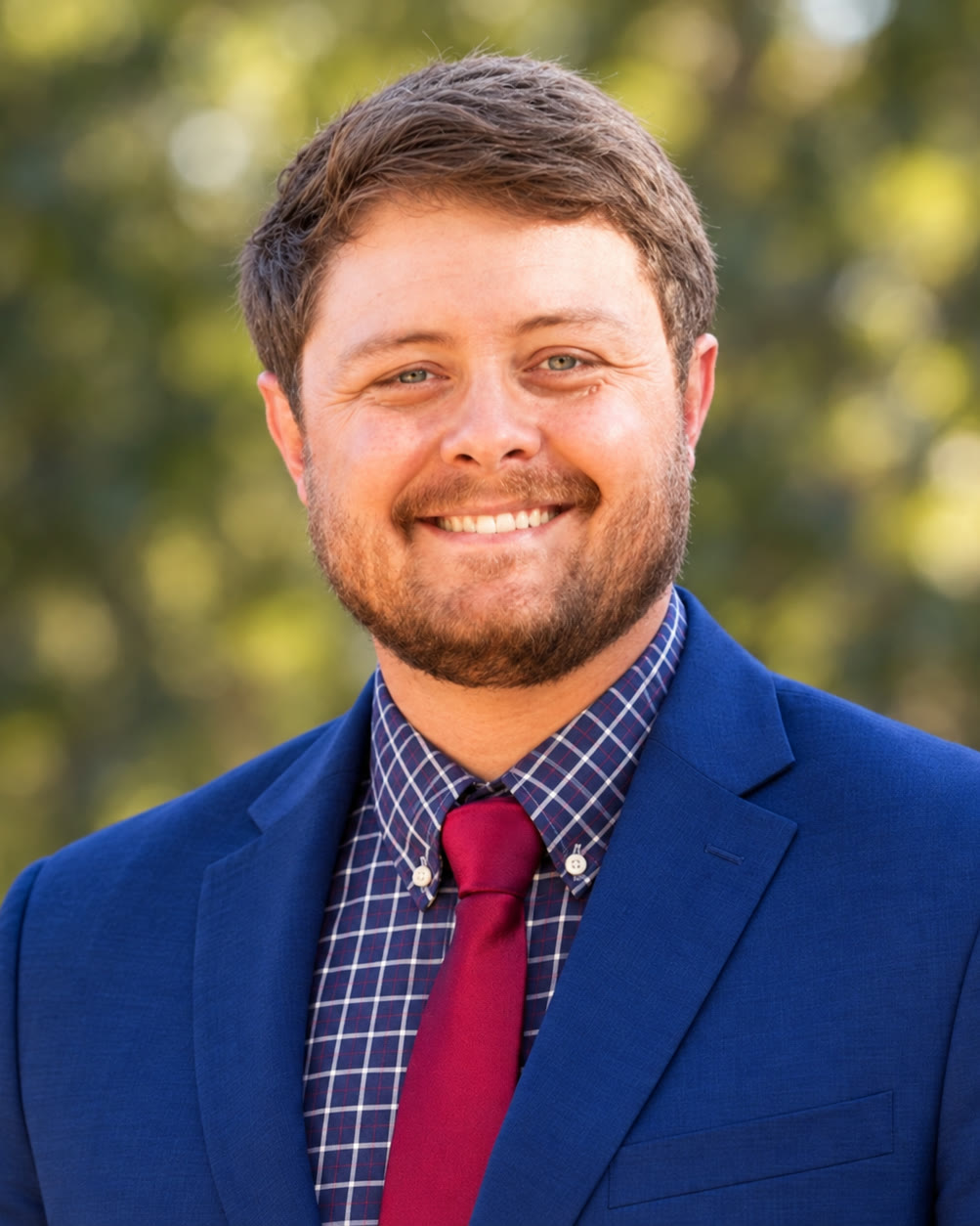 Justin Gray smiling outdoors in a navy blazer, checked shirt and red tie
