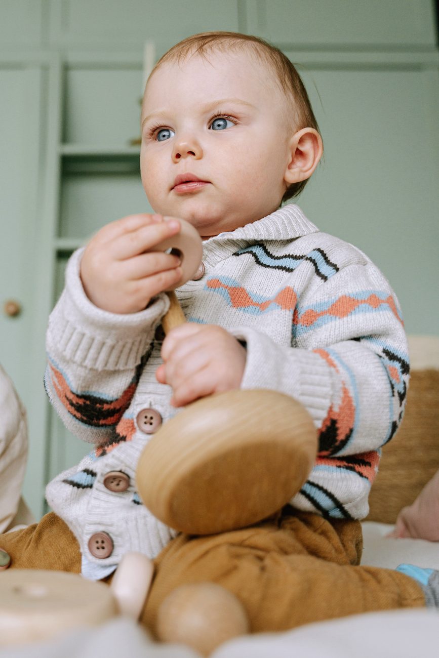 A baby in a knit sweater holding a wooden rattle